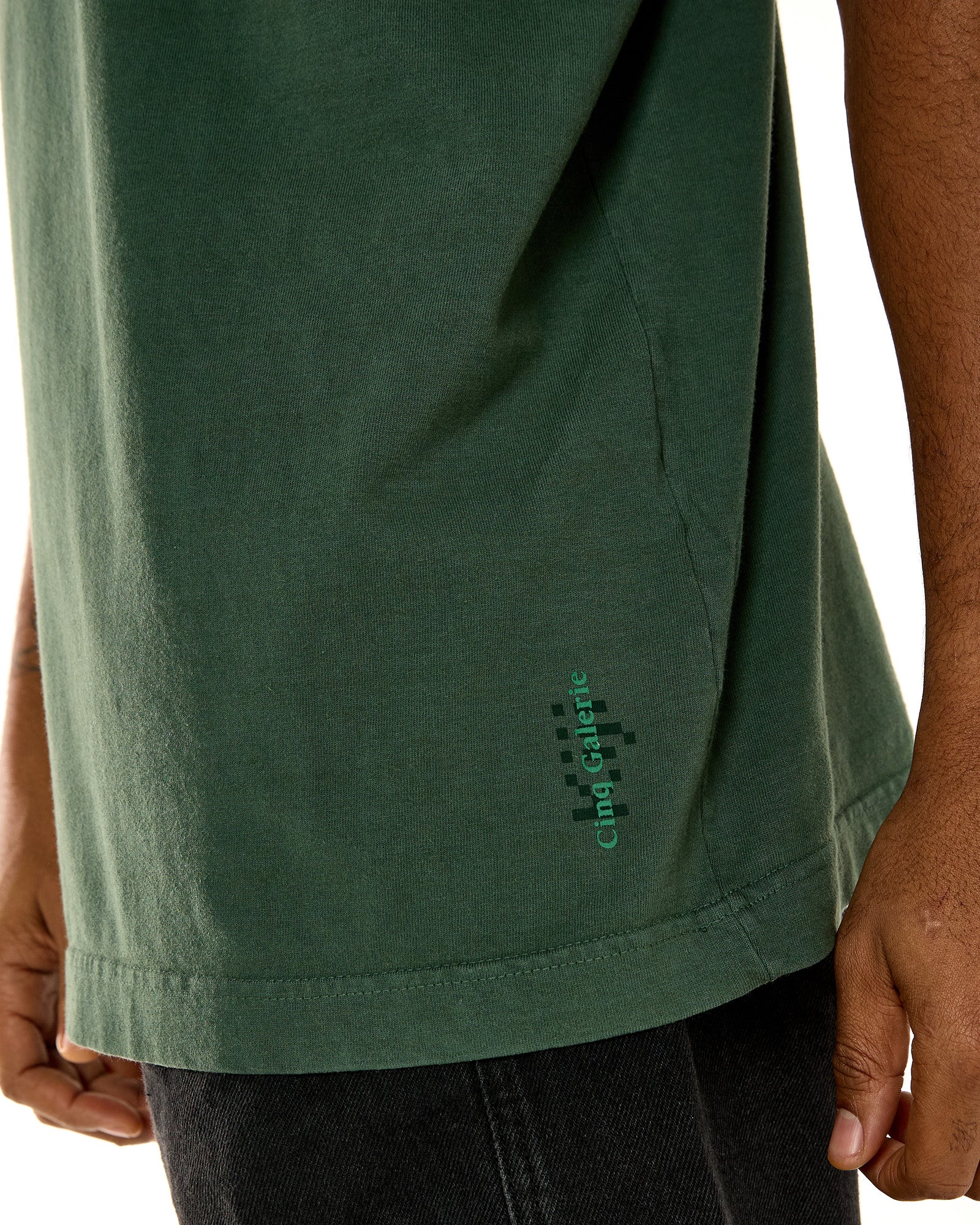 Detalhe do silk relevo da Kiji em colaboração com a cinq galerie na lateral esquerda da camiseta. o silk conta com o logo da KIJI em um tom de verde mais escuro do que a camiseta, e acima dele, há o logo da cinq, também em relevo, porém em um tom de verde mais claro do que a camiseta.