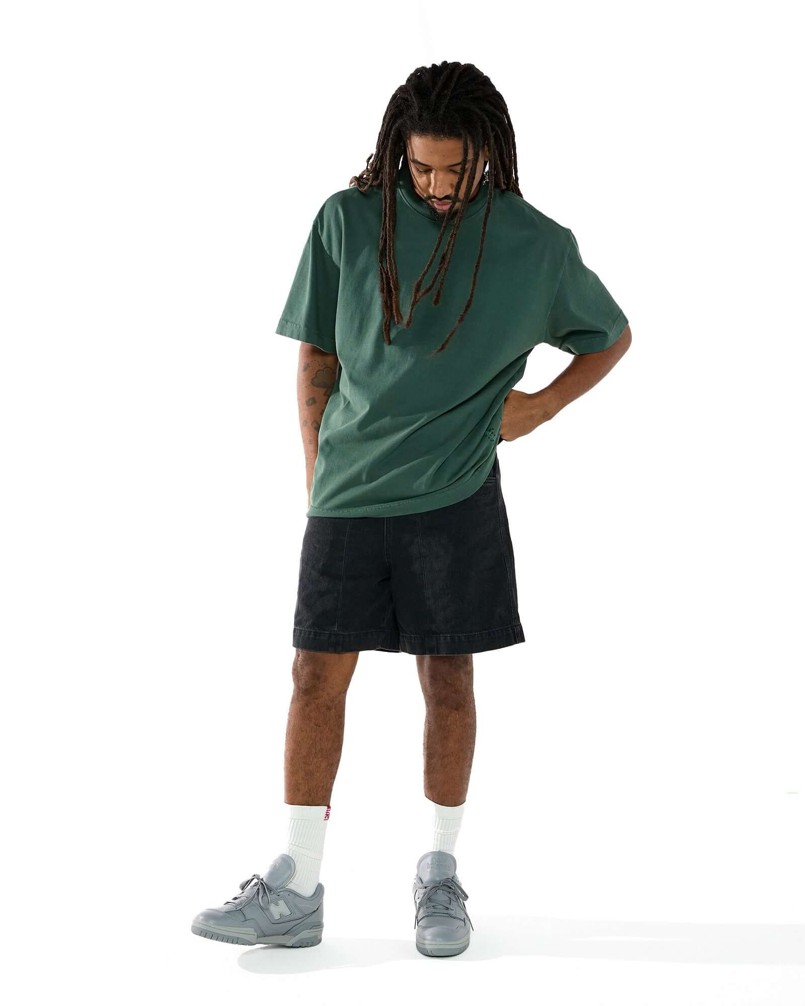 Modelo vestindo a camiseta oversized off white Kiji, uma bermuda jeans preta, meia flag branca, e um tênis cinza.