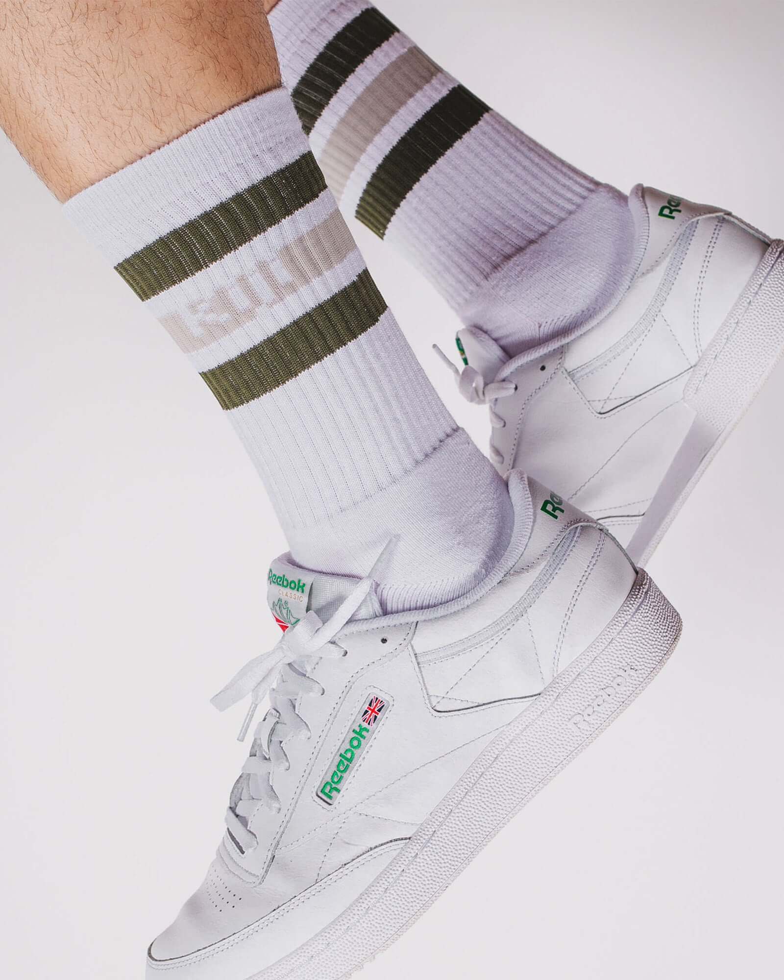 Foto de detalhe, zoom no pé do modelo vestindo a meia Kiji Stripes SK8 de algodão e um tênis branco.