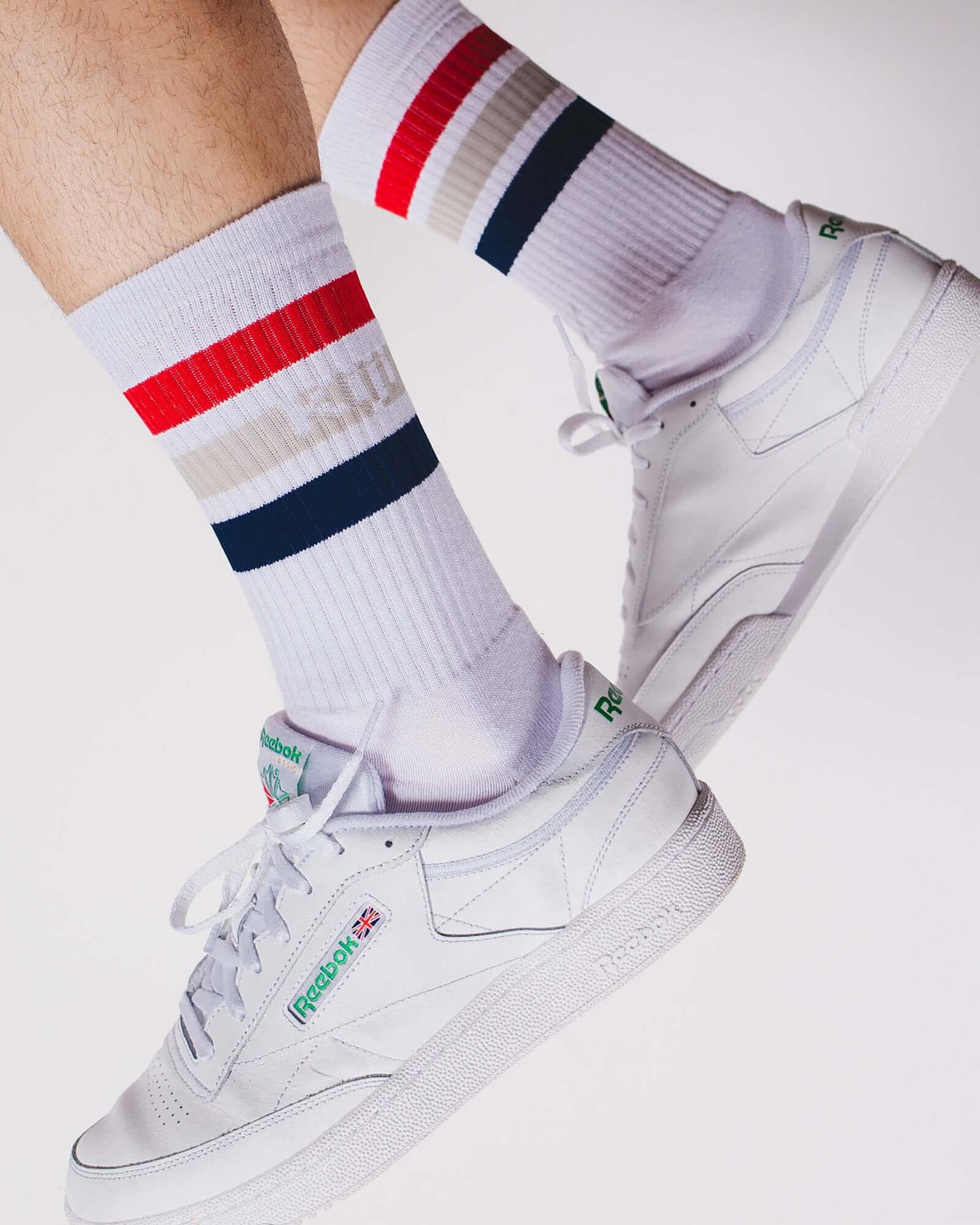 Foto de detalhe, zoom no pé do modelo vestindo a meia Kiji Stripes SK8 de algodão e um tênis branco.