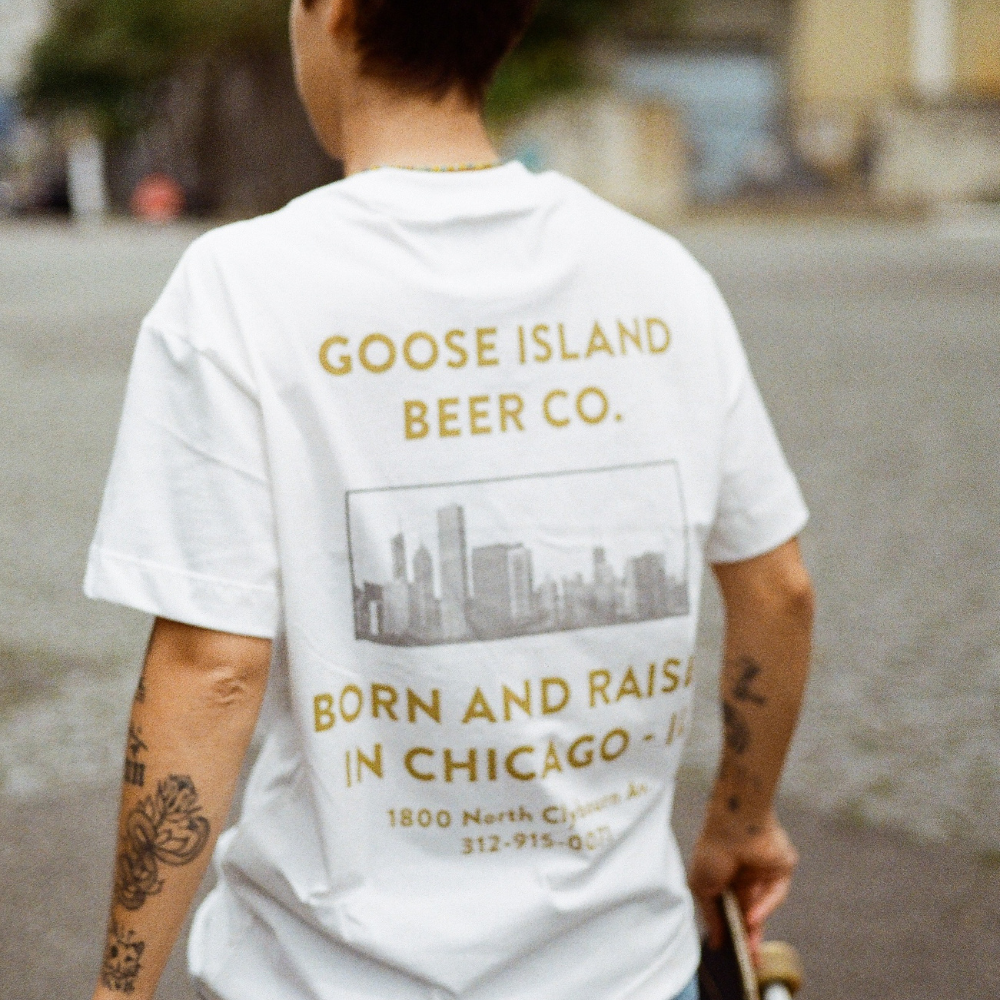 Foto de uma modelo de costas segurando um skate e usando a T-shirt Classic Kiji x Goose Island Born and Raised Branca