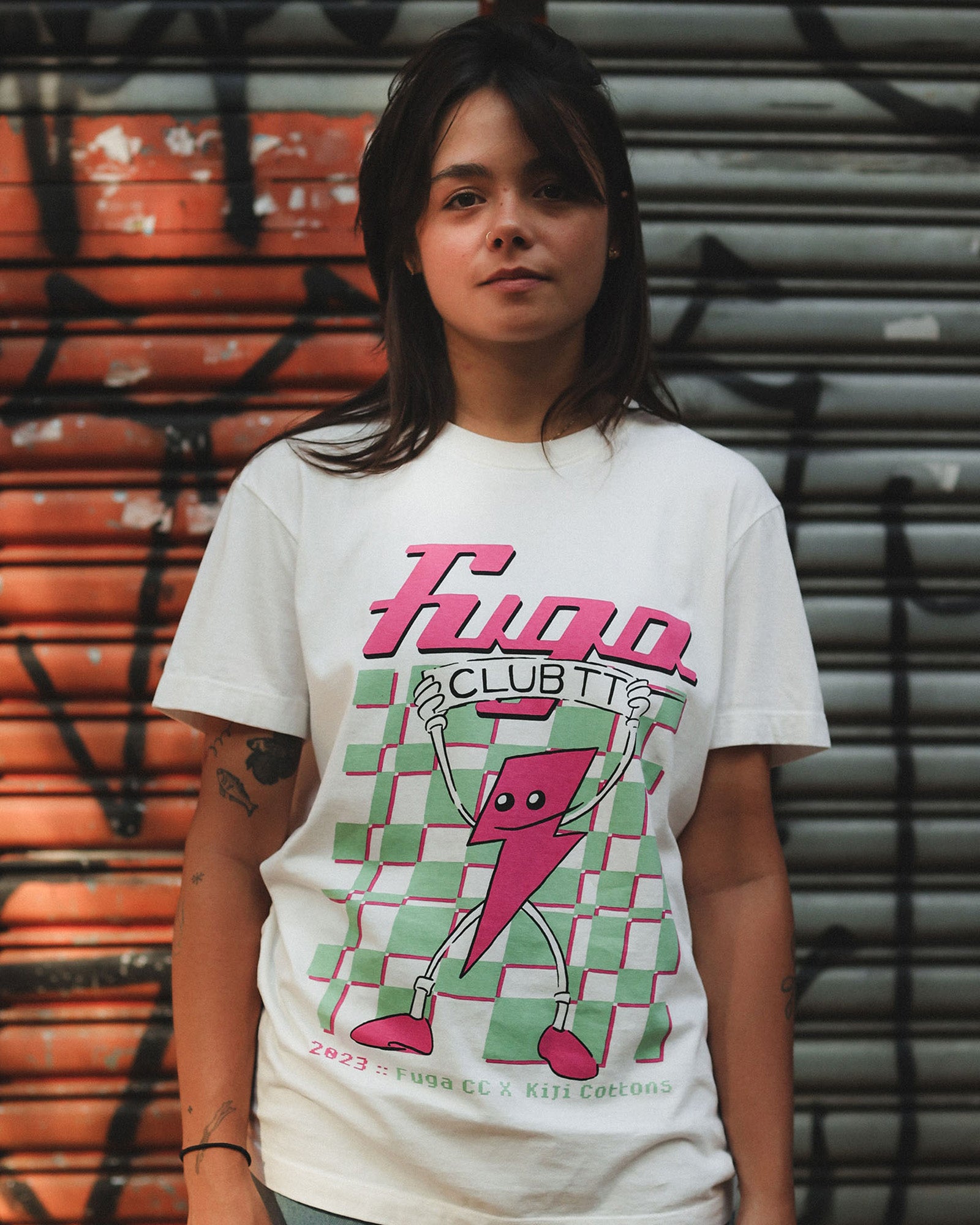 Modelo vestindo a T-Shirt Classic Kiji Off-White TT23. A estampa da colaboração o FugaCC conta com o Mascote do clube (Um raio na cor rosa) segurando uma faixa escrito ClubTT, logo acima há o logo do Fuga e atrás uma malha quadricula estilizada em verde, com sombras rosas. Já na parte debaixo, está escrito 2023 e Fuga CC x Kiji Cottons em uma fonte inspirada na estética 8-bit