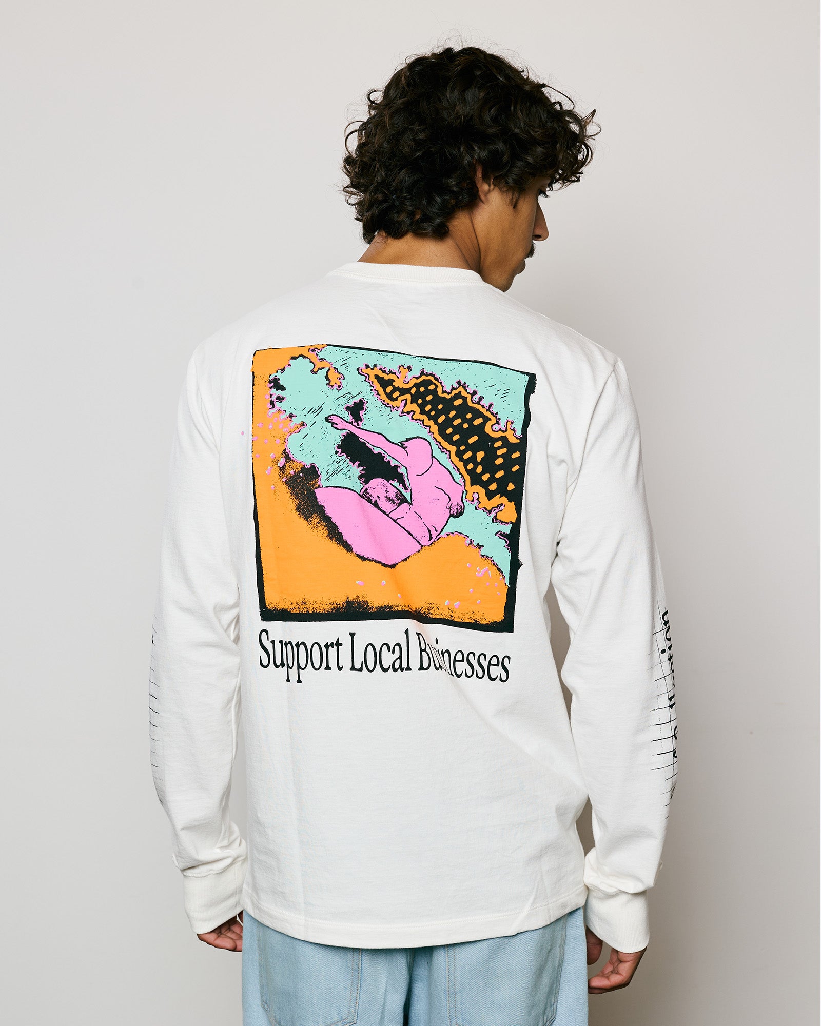 Modelo usando camiseta de manga longa branca com estampa “Support Local Businesses” nas costas.