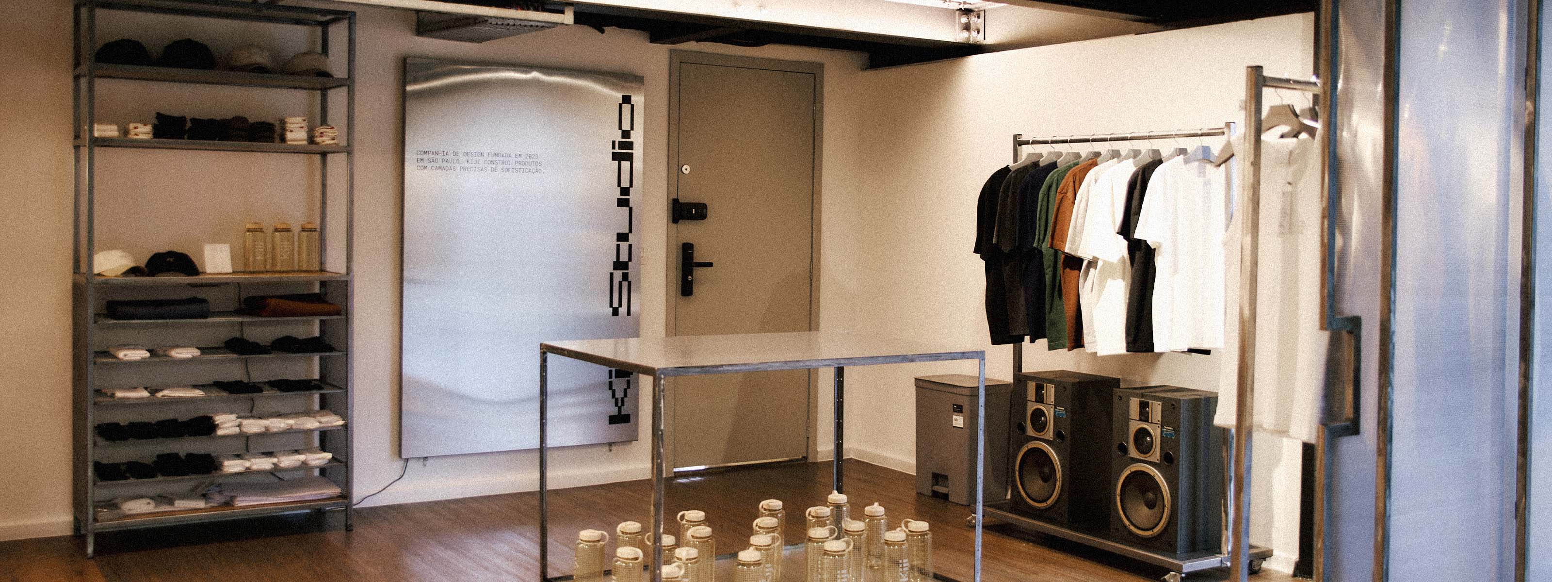 Interior do estúdio/loja física da Kiji em São Paulo, com estante metálica de bonés, camisetas dobradas e meias à esquerda, arara de camisetas à direita ao lado de duas caixas de som vintage, e um quero de aço inox com o texto 'Companhia de design fundada em 2023 em São Paulo, Kiji constrói produtos com camadas precisas de sofisticação' e o logo 'Kiji Studio' na parede central.